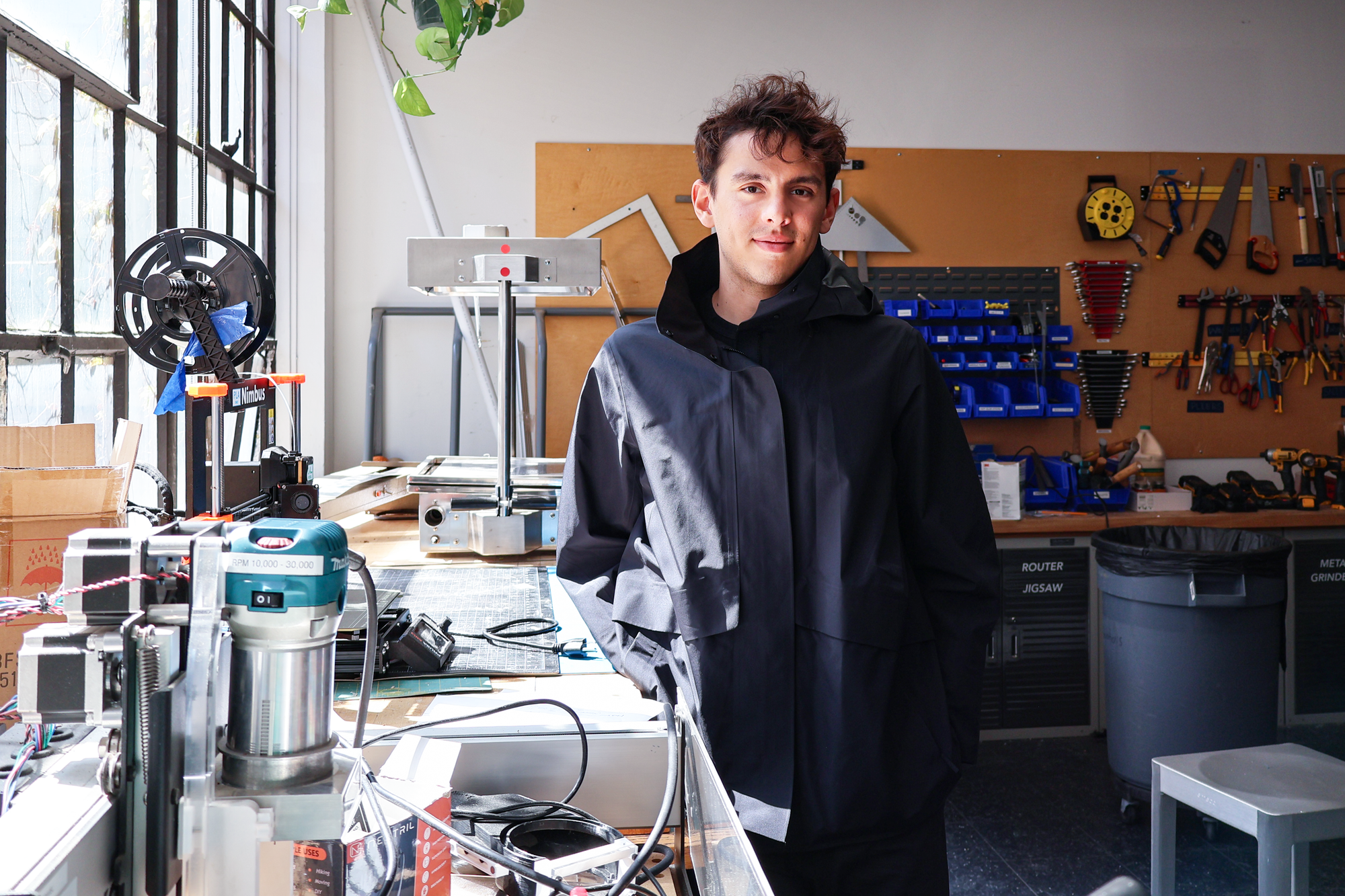 Young man standing in a sunlit makerspace or workshop, wearing a black jacket. He is surrounded by tools and equipment, including a router, spools of filament, and various hand tools mounted on a pegboard behind him. Large windows let in natural light.